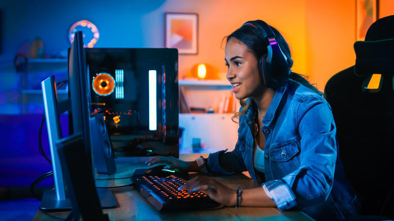 Jolie et excitée Black Gamer Girl in Headphones joue à un jeu vidéo en ligne à la première personne sur son ordinateur.  La chambre et le PC ont des lumières LED au néon colorées.  Soirée confortable à la maison.