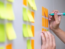 IT worker tracking his tasks on kanban board. Using task control of agile development methodology. Man attaching sticky note to scrum task board in the office.
