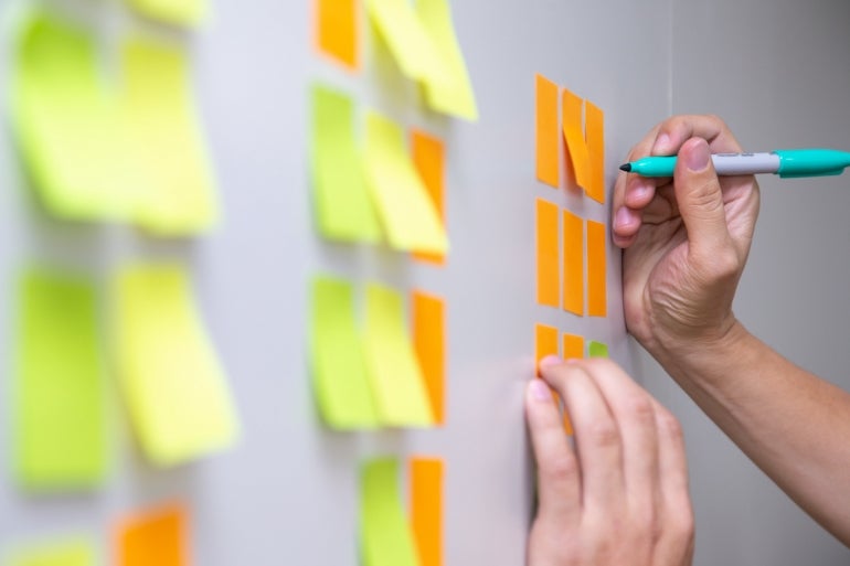 IT worker tracking his tasks on kanban board. Using task control of agile development methodology. Man attaching sticky note to scrum task board in the office.