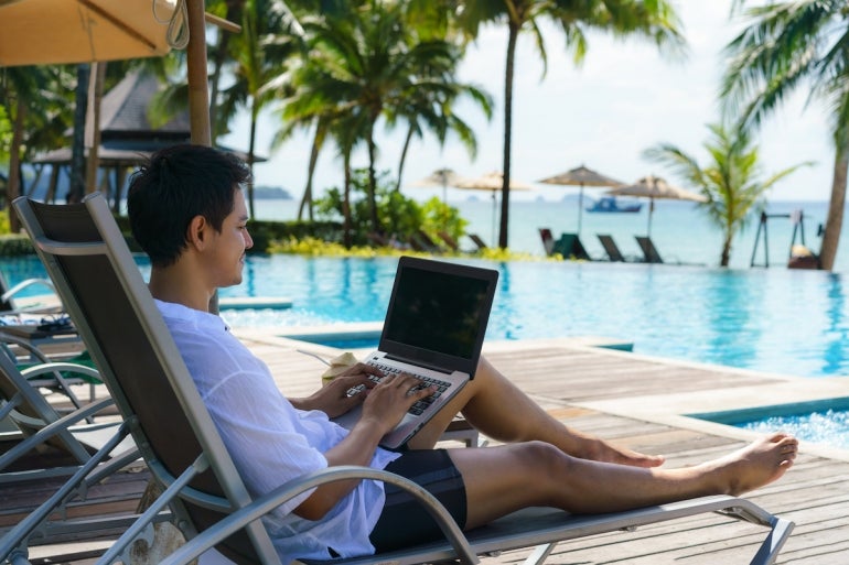 Asian man spent his summer vacation working on his laptop in a chair near the swimming pool in resort hotel near sea.