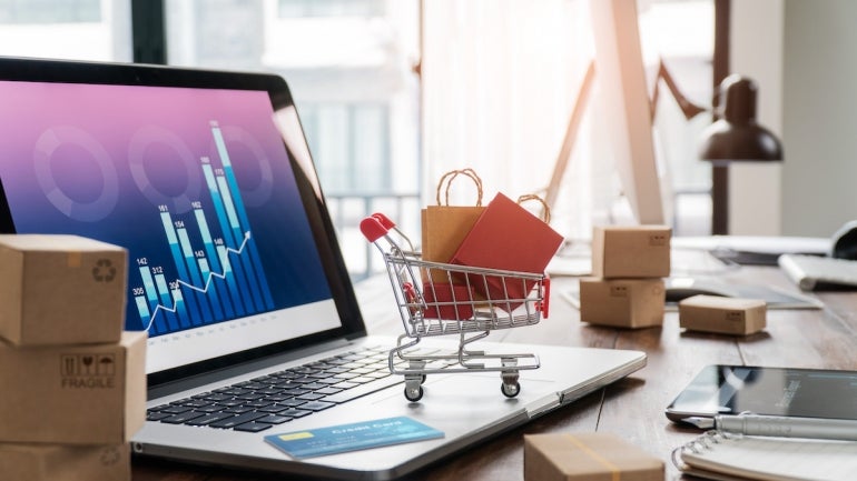 Shopping bags in shopping cart and credit card on laptop with paper boxes on table and sales data economic growth graph on screen, buying and selling services online on network, online shopping and e-commerce concept.