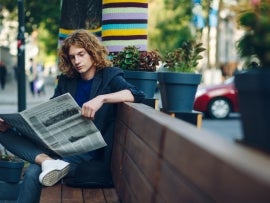 Red haired hipster man sitting on bench reading newspaper
