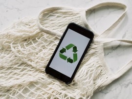 Smartphone with recycling sign on screen placed on white mesh bag on marble table.