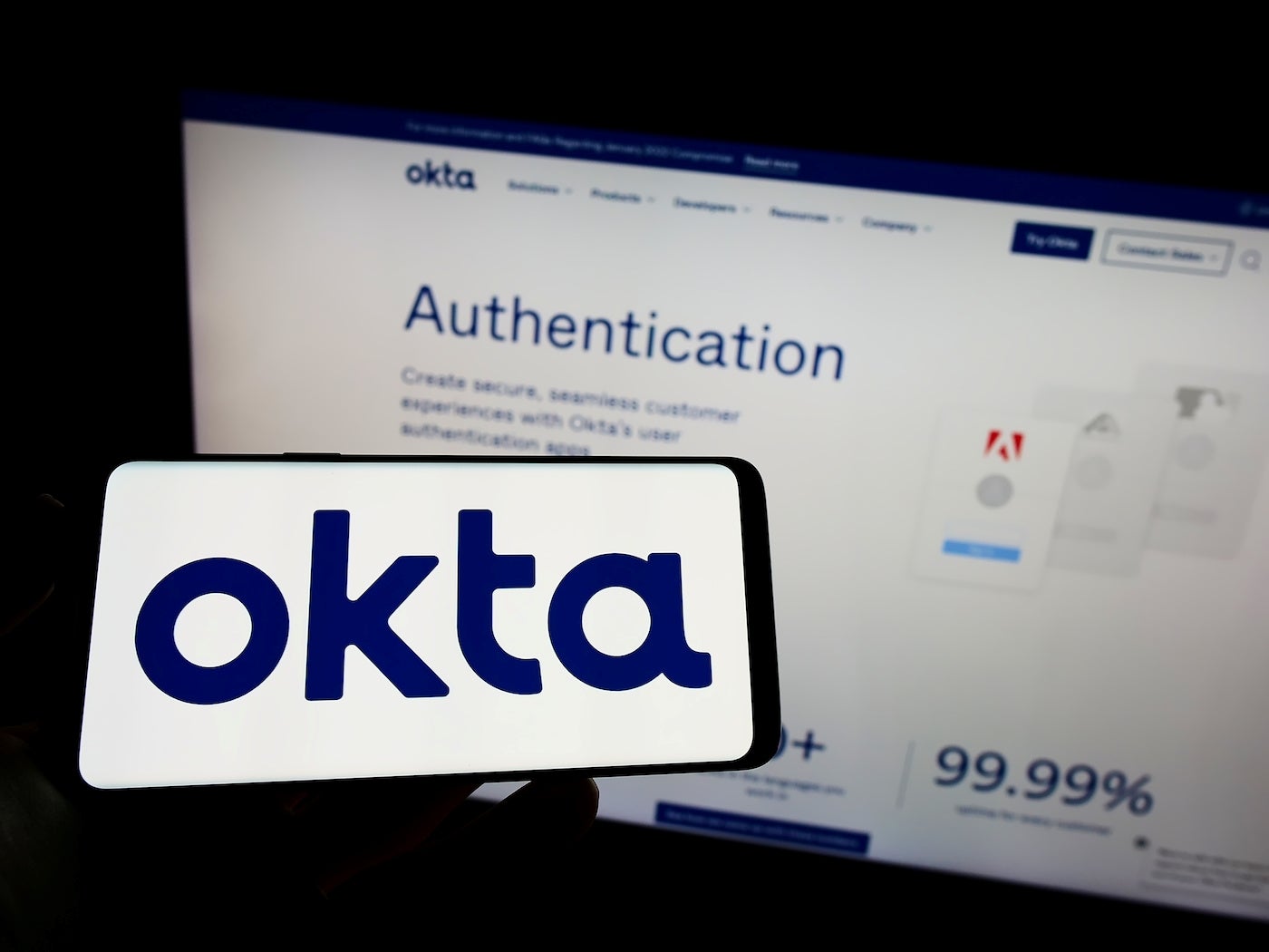 A person holding up their phone with the Okta logo on it in front of a page on the Okta website for Authentication
