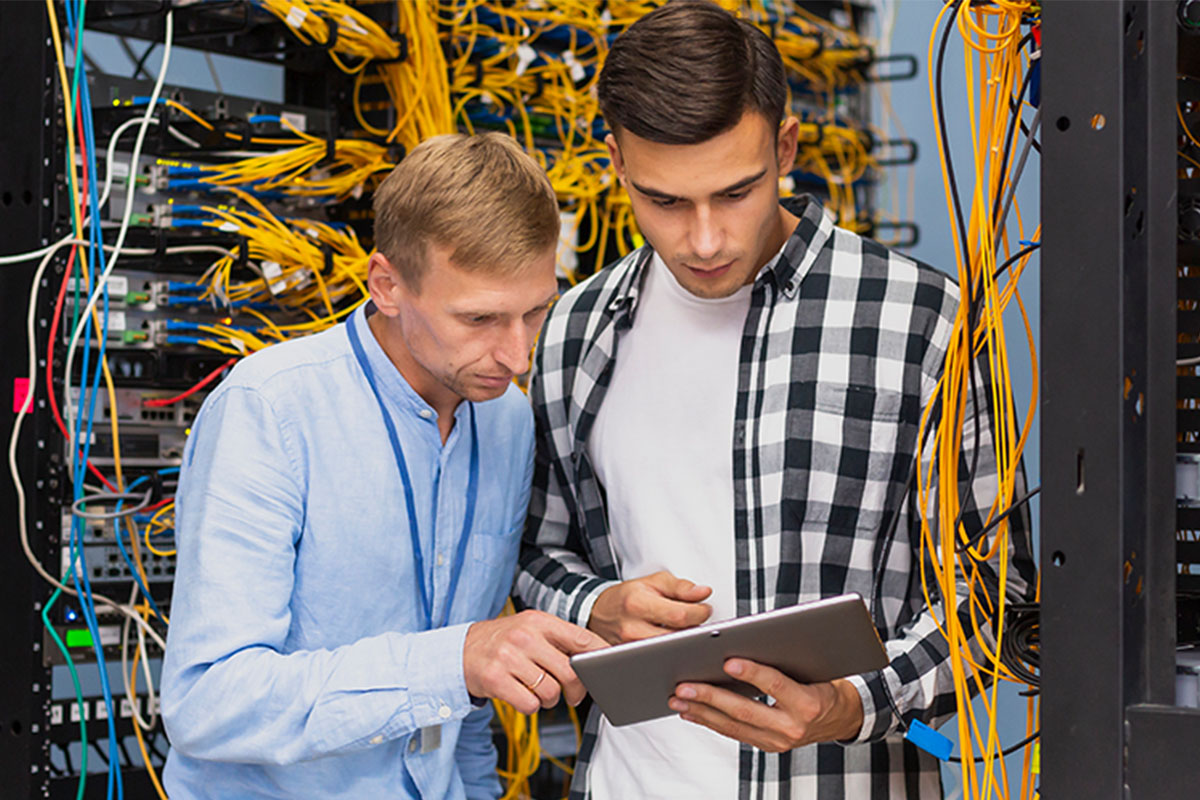 two guys standing in front of a bunch of server cables looking at a tablet together