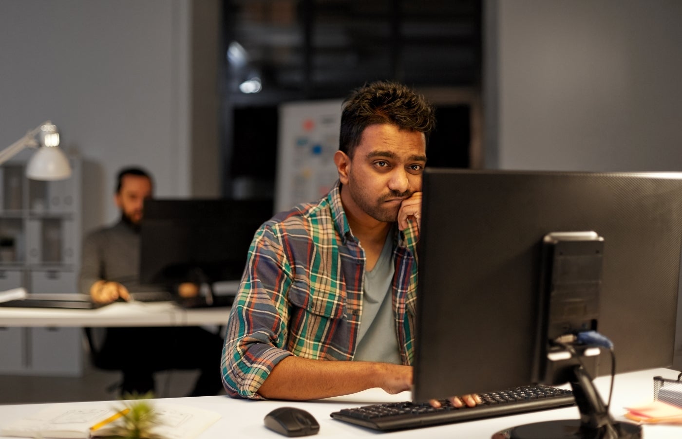 A visibly tired/frustrated man working at a computer in an office setting with another coworker in the background
