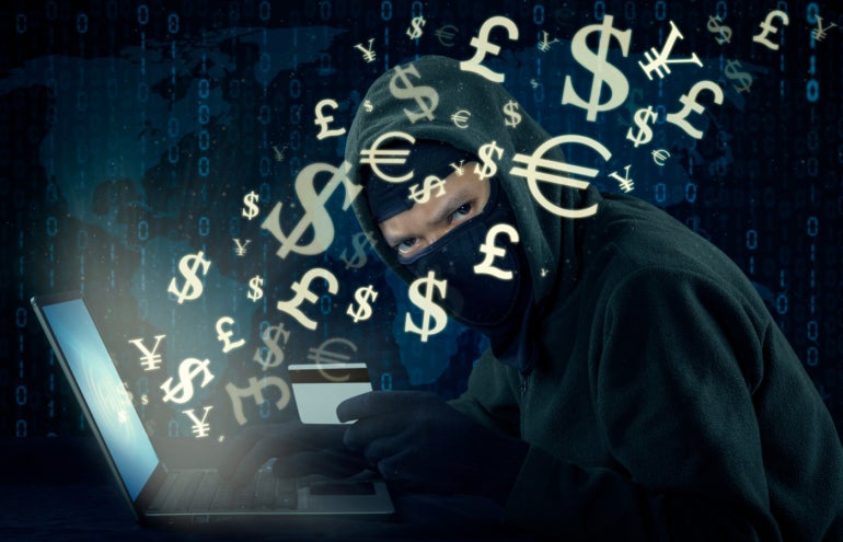 Image of male hacker wearing mask and using laptop while holding credit card to steal money through online transaction.